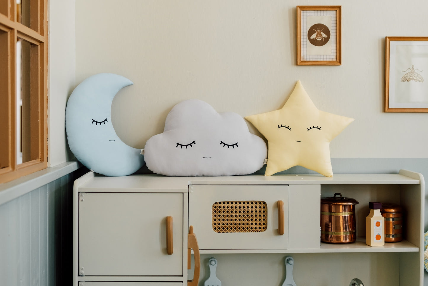 Decorative pillows shaped like a moon, cloud, and star on a shelf with framed pictures on the wall.
