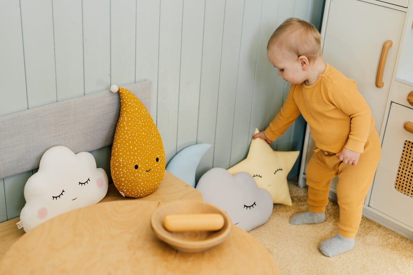 Baby in a mustard yellow outfit playing with colorful cushions in a nursery.