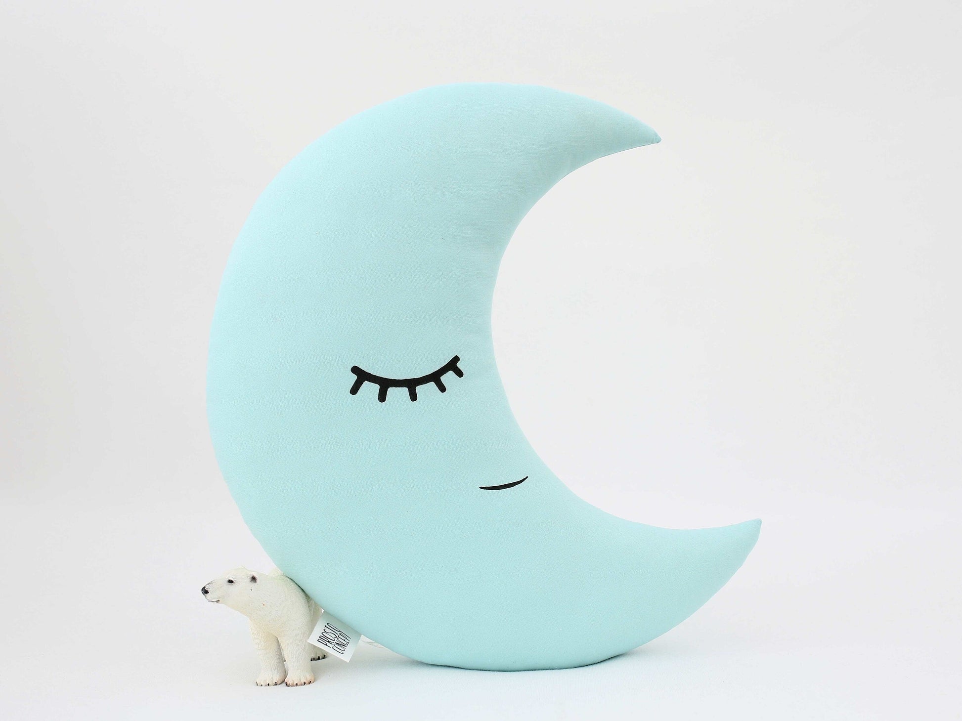 Mint crescent moon throw pillow, sleeping face and small polar bear toy at base – upright in nursery setting.