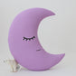 Lavender crescent moon throw pillow, simple sleeping face design – plain surface, small polar bear figurine partly visible underneath.
