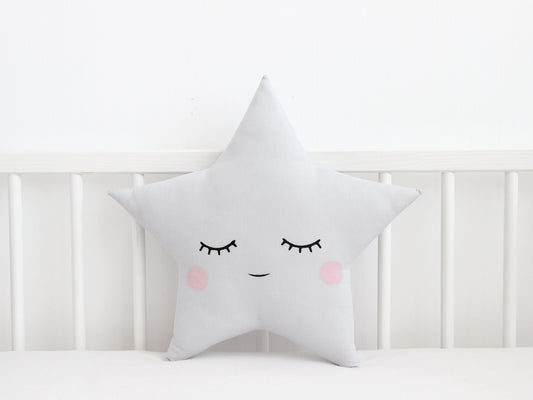 Light gray star throw pillow, smiling face with closed eyes – resting against white crib rail on plain white background.